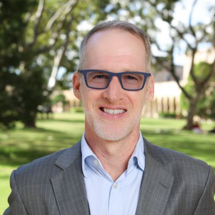 Andrew Flannery stands in the foreground with greenery and sandstone buildings behind him.